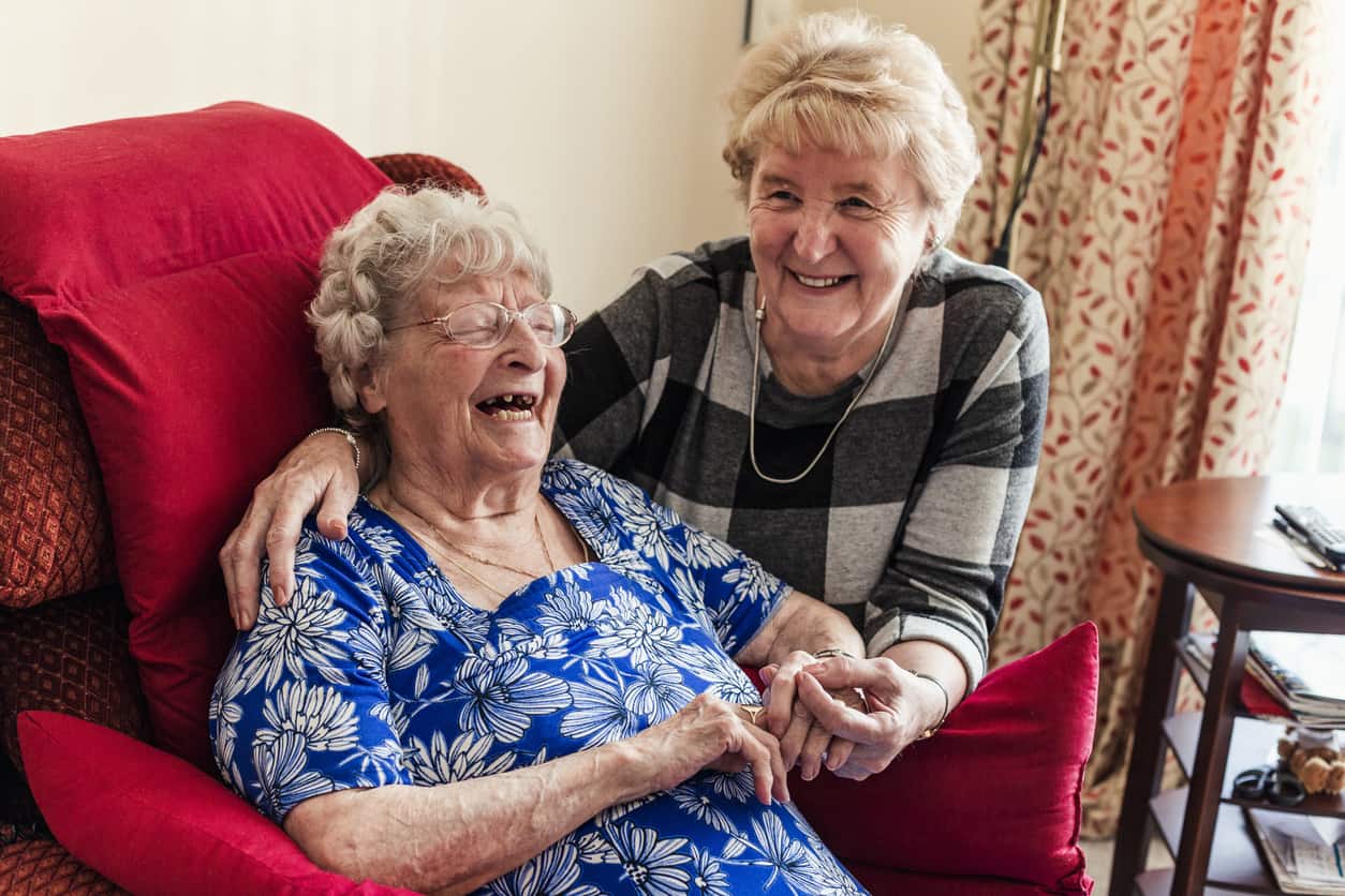 One elderly lady sits with her arm around another with loving care. They are in a nursing home in Newcastle upon Tyne, North East England.
